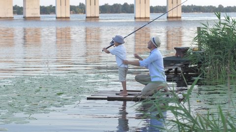 family hobby, male fisherman teaches little boy to fish on fishing rod sitting on wooden raft in a river among reeds