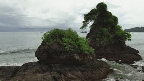 Aerial panning view of tropical shoreline, rocks in the ocean, overgrown with vegetation, dense rainforest in the background, wild nature and force of elements