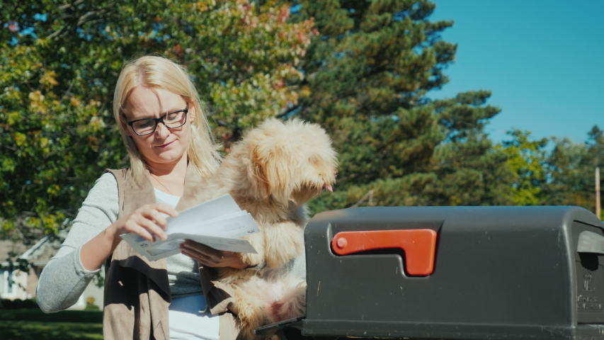 A young woman with a small dog in her arms picks up letters from a mailbox
