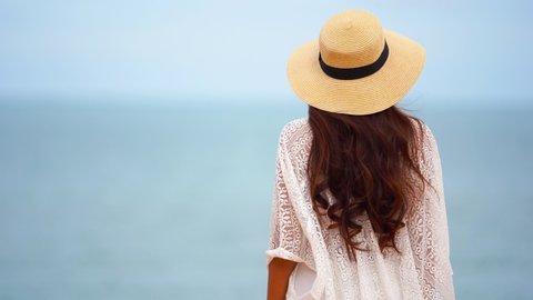 Woman with long brown hair, a lacey white beach coverup, and floppy straw? hat walks toward the incoming tide.