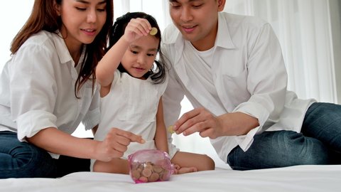 Asian family happy in the house. Father, mother and daughter aged three putting coin into piggy bank for saving on the bed. Happiness family savings concept. slow motion