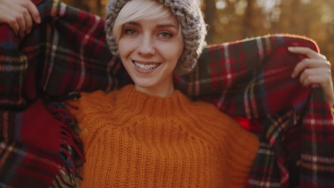 Young happy smiling tourist girl with orthodontic braces on her teeth, wears, wraps in red tartan scarf, enjoys sunny autumn in forest, park
