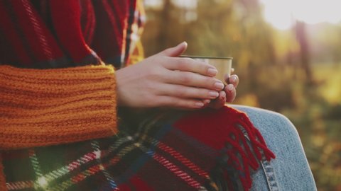 Close up of woman`s hands holding cup of hot tea. Tourist girl wearing tartan scarf enjoys warm drink in autumn park