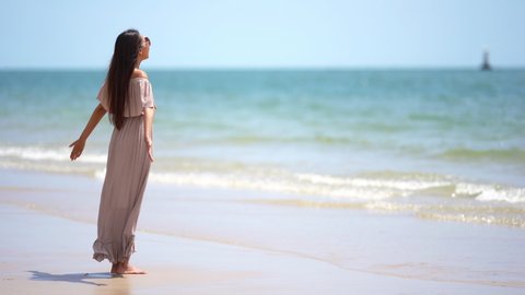 Attractive Asian woman standing on the beach with arms spread as wind blows across her body and small gentle waves of turquoise water wash onto the sand. Bright tropical sunlight and island vacation.