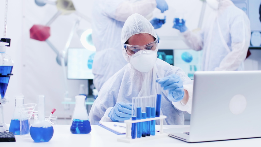Female scientist picking up a test tube with blue solution and typing on computer. Scientist in protection uniform. | Shutterstock HD Video #1035602378