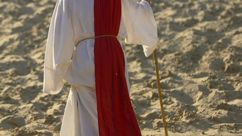 Jesus Christ in robe and red sash walking through desert, looking at camera