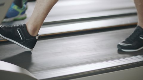 Closeup feet walking on treadmill in fitness gym slowly. Black sneakers training on running machine. Fit legs jogging in sport club in slow motion.