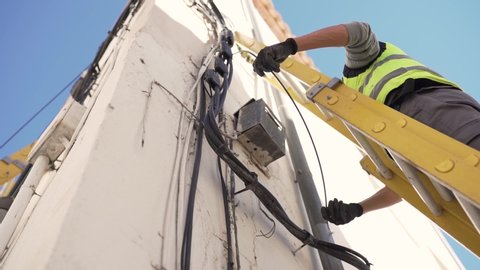 Low Upwards shot or a technician in protective gear on a ladder installing optical fiber cables on a building