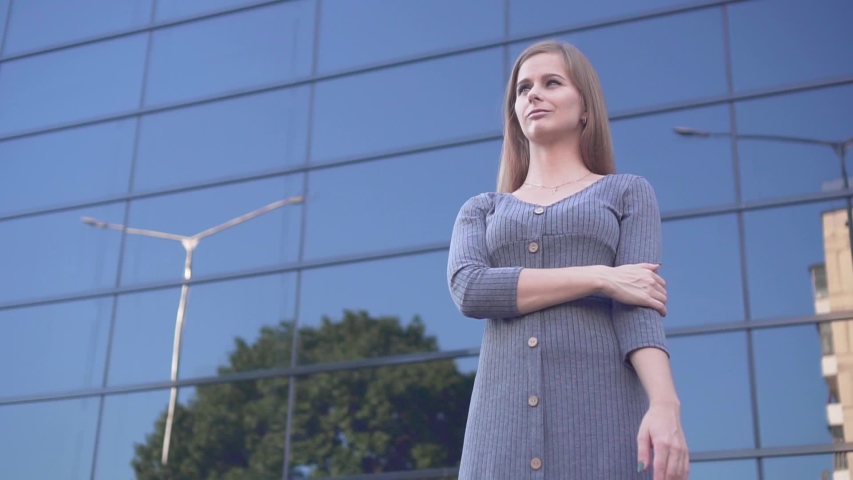 Cute young girl walks in an urban environment. Happy girl in a gray dress near a modern glass building.