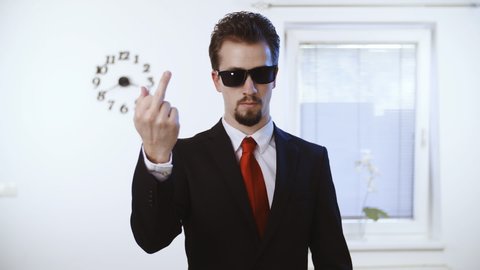 Businessman with glasses shows the fuck you middle finger. Medium shot of a male person in focus dressed up nicely with a red tie. Wall with clock and window in the background.