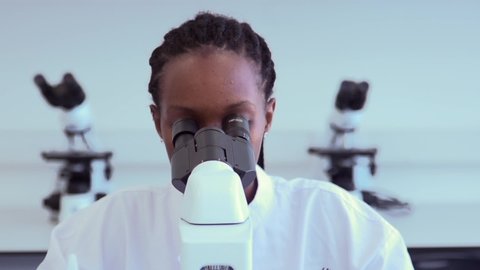 African-American scientist researching in laboratory, looking through the microscope and taking notes