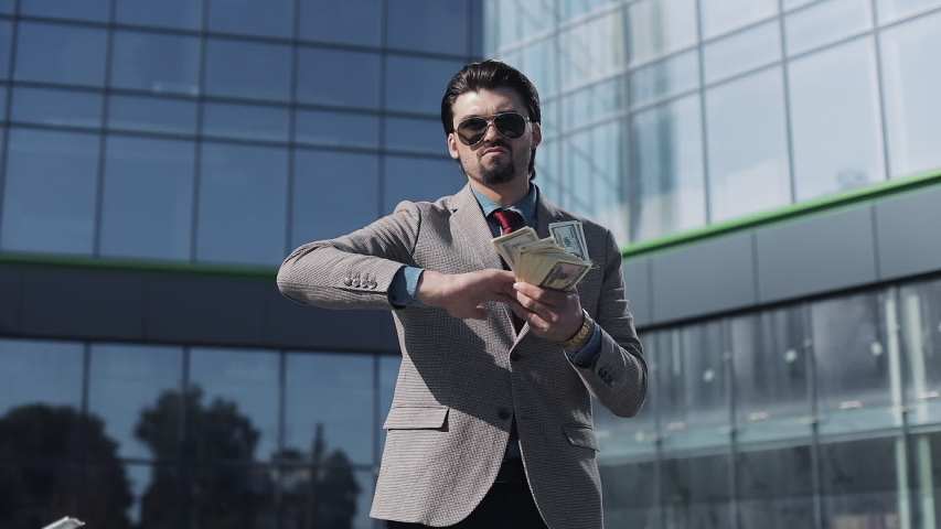 Young Successful Businessman in a Suit Wear Sunglasses Throwing Money in the Camera Standing in the Street near Office Building. Money rain, falling dollars. Successful business concept. Royalty-Free Stock Footage #1035651386