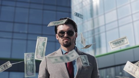 Young Successful Businessman in a Suit Wear Sunglasses Throwing Money in the Camera Standing in the Street near Office Building. Money rain, falling dollars. Successful business concept.