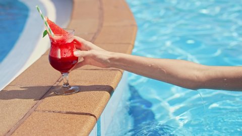 Close-up: the female hand takes a cool cocktail from the pool. The girl takes a soft drink red, swimming in the pool. Summer day, blue water, bright, juicy lemonade. Slow motion, 4K