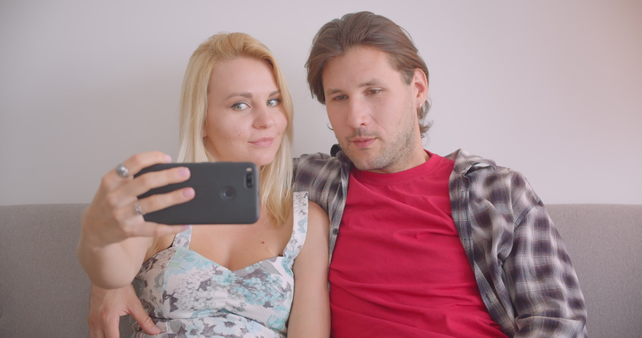 Closeup portrait of adult attractive caucasian couple taking selfies on phone smiling happily sitting on couch indoors in apartment