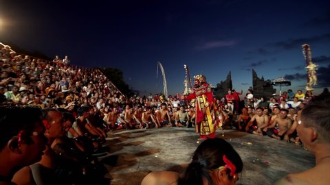 BALI, INDONESIA - JUNE 10, 2019: Outdoor theater full of viewers and stage with actor in mask performing traditional Kecak dance. White beast on the stage. Uluwatu temple, evening