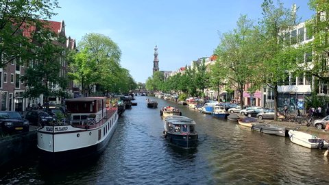 Amsterdam / Netherlands - 06 22 2019: Boats drive through the banks under the bridges in good weather. In the background the church of Amsterdam Netherlands