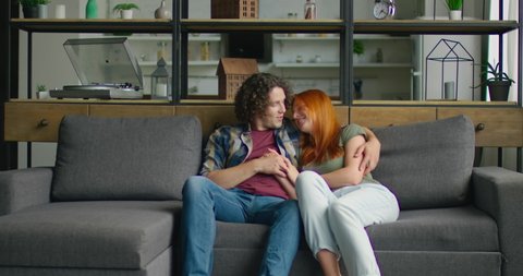 Young couple in love is sitting on sofa at home, in romantic mood, hugging, being happy, enjoying their relationships, man makes compliments to woman, Caucasian. 4K, shot on RED camera.