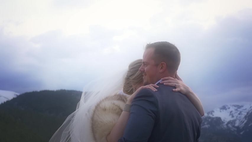 A bride and groom sway forehead to forehead in the mountains, her veil gently blowing up into the wind