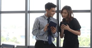 Portrait scene video of handsome Caucasian businessman using tablet computer and beautiful Asian businesswoman using smartphone with happy mood in break time in office.
