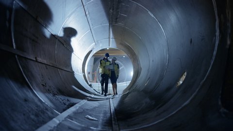Two Heavy Industry Engineers Walking Inside Pipe, Use Laptop, Have Discussion, Checking Welding. Construction of the Oil, Natural Gas and fuels Transport Pipeline. Industrial Manufacturing Factory