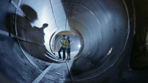 Two Heavy Industry Engineers Walking Inside Pipe, Use Laptop, Have Discussion, Checking Design. Construction of the Oil, Natural Gas and fuels Transport Pipeline. Industrial Manufacturing Factory