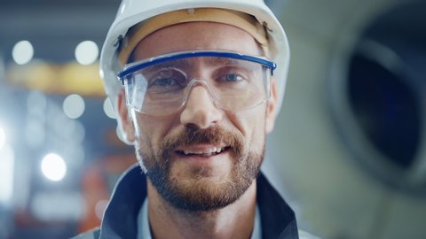 Portrait of Smiling Professional Heavy Industry Engineer / Worker Wearing Safety Uniform, Goggles and Hard Hat. In the Background Unfocused Large Industrial Factory