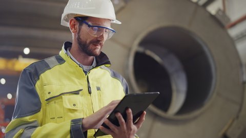 Portrait of Professional Heavy Industry Engineer / Worker Wearing Safety Uniform and Hard Hat Uses Tablet Computer. In the Background Construction Factory for Oil, Gas and Fuels Transport Pipeline