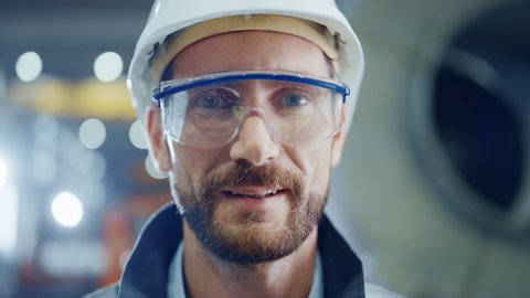 Portrait of Smiling Professional Heavy Industry Engineer / Worker Wearing Safety Uniform, Goggles and Hard Hat. In the Background Unfocused Large Industrial Factory