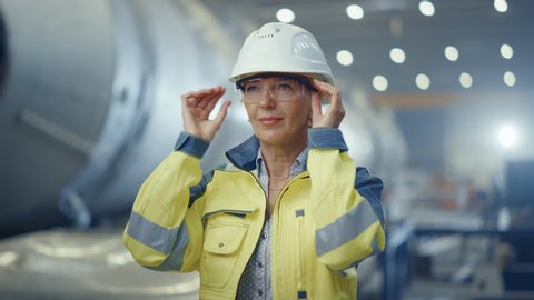 Portrait of Professional Heavy Industry Female Engineer Wearing Safety Uniform and Hard Hat, Taking off Goggles. In the Background Unfocused Large Industrial Factory where Welding Sparks Flying
