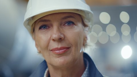 Portrait of Professional Heavy Industry Female Engineer Wearing Safety Uniform and Hard Hat, Smiling Charmingly. In the Background Unfocused Large Industrial Factory where Welding Sparks Flying