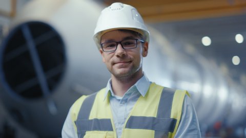 Portrait of Young Professional Heavy Industry Engineer / Worker Wearing Safety Vest, Putting on Hardhat. In the Background Unfocused Large Industrial Factory where Welding Sparks Flying. Slow Motion