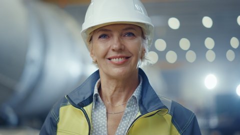 Portrait of Professional Heavy Industry Female Engineer Wearing Safety Uniform and Hard Hat, Smiling Charmingly. In the Background Unfocused Large Industrial Factory where Welding Sparks Flying