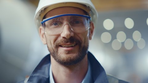 Portrait of Professional Heavy Industry Engineer / Worker Wearing Safety Uniform, Goggles and Hard Hat Smiling. In the Background Unfocused Large Industrial Factory where Welding Sparks Flying