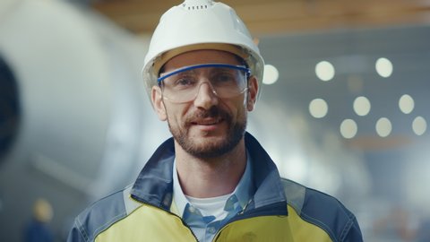 Portrait of Professional Heavy Industry Engineer / Worker Wearing Safety Uniform, Goggles and Hard Hat Smiling. In the Background Unfocused Large Industrial Factory where Welding Sparks Flying