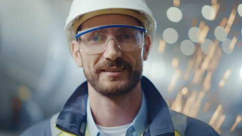Portrait of Smiling Professional Heavy Industry Engineer / Worker Wearing Safety Uniform, Goggles and Hard Hat. In the Background Unfocused Large Industrial Factory where Welding Sparks Flying