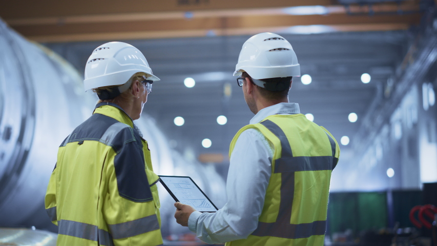 Two Heavy Industry Engineers Stand in Pipe Manufacturing Factory, Use Digital Tablet Computer, Have Discussion. Construction of Oil, Gas and Fuels Transport Pipeline. Back View Sparks Flying | Shutterstock HD Video #1035704123