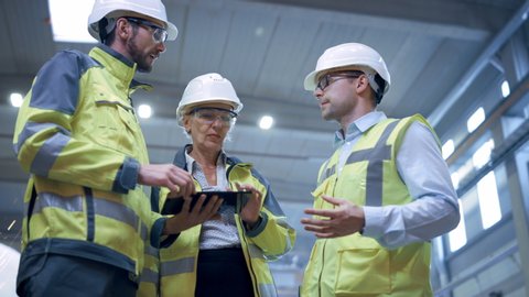 Three Heavy Industry Engineers Stand in Pipe Manufacturing Factory, Use Digital Tablet Computer, Have Discussion. Design and Construction of Large Oil, Gas and Fuels Transport Pipeline. Low Angle Arc