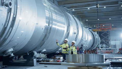 Two Heavy Industry Engineers Walking Through Pipe Manufacturing Facility, Use Digital Tablet, Have Discussion. Modern Industrial Design and Construction of Oil, Gas and Fuels Transport Pipeline
