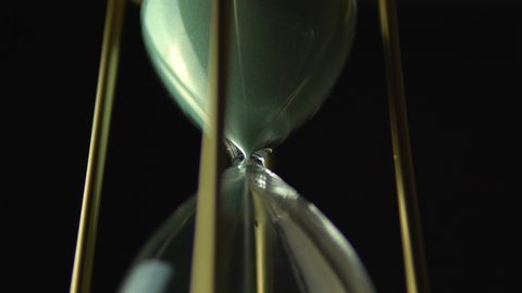 Slow motion rotation of sand timer clock with sand flowing from through hour glass. View from below hourglass close up macro shot. Black background.