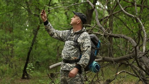 young handsome caucasian man in a camouflage suit, cap and backpack takes selfie for social networks in a beautiful dense green forest, close-up