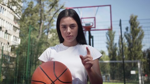Portrait cute brunette woman with a basketball ball challenging viewer pointing her finger at the camera. Concept of sport, power, competition, active lifestyle. Sports and recreation.