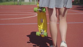 Legs of woman in short skirt with a yellow skateboard walking on the basketball court. Shadow follows the girl. Concept of sport, power, competition, active lifestyle
