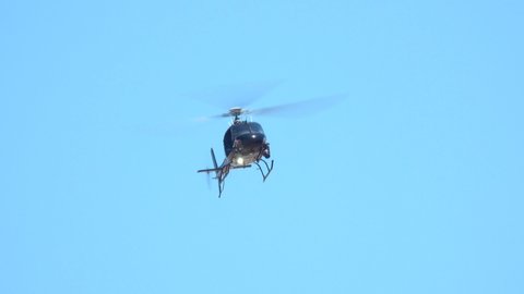 A modern, black helicopter hovers in the sky, as viewed from the ground, with the front of the aircraft facing the viewer.