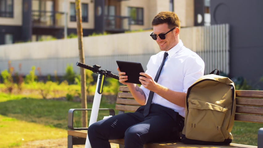 business, technology and people concept - young smiling businessman with tablet computer, backpack and electric scooter sitting on street bench in city