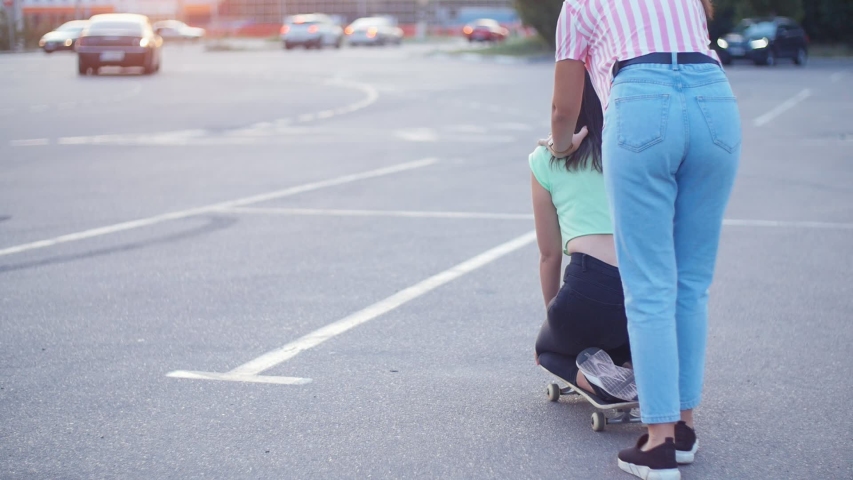 Positive girls having fun with scateboard on the parking at sunset in Slow motion. Two young cheerful women riding on the skateboard