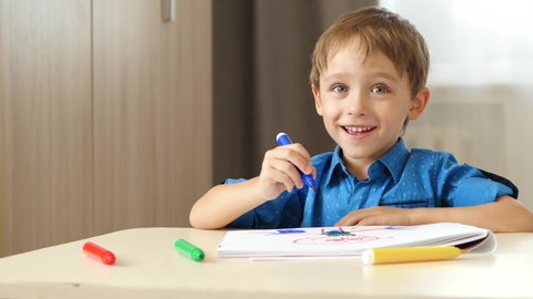 Portrait of a happy boy caucasian appearance, who sits at the table and draws felt pen in the album. The child looks at the camera and laugh.