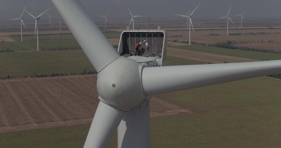 Drone flies in a circle from right to left to two workers who work on a huge windmill. Workers repair a windmill at an altitude during sunny weather. Many windmills spin in the background. Grey sky | Shutterstock HD Video #1035763067