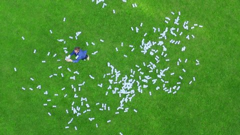 A young man businessman or student or office worker in a blue checkered jacket runs on a green grassy lawn strewn with money and throws 100-dollar bills. Jackpot winners concept. Aerial view.