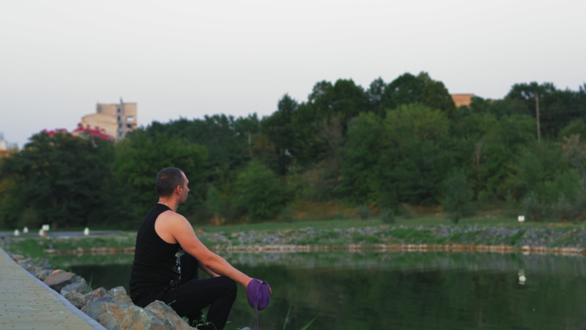 Man Sits on Stones on Lake Shore and Repeats Mantra Holding Pouch with Rosary in Hand